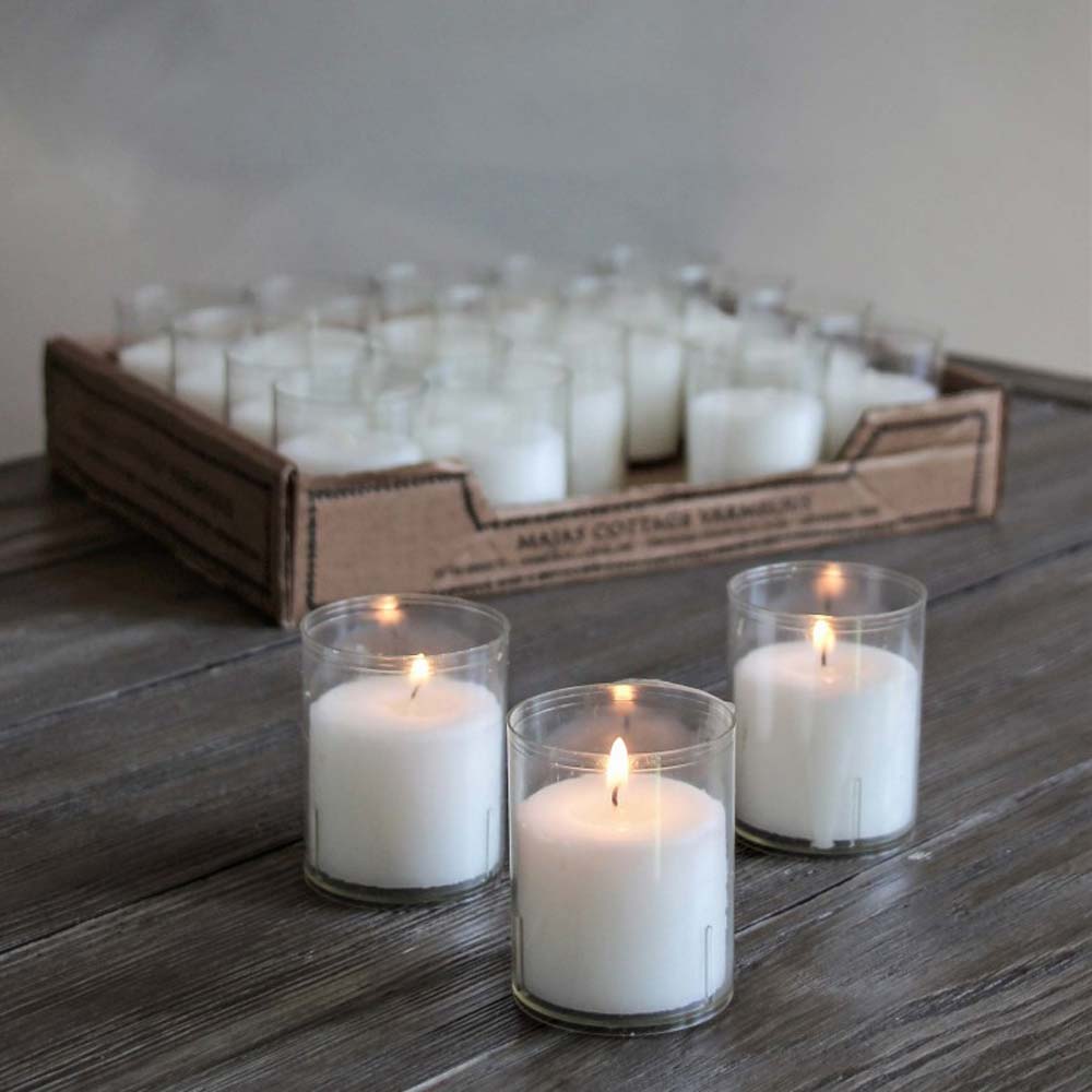Eine Sammlung kleiner brennender Kerzen in durchsichtigen Glashaltern, ausgestellt auf einer Holzoberfläche, mit einem Karton mit der Aufschrift „Majas Cottage - Teelicht