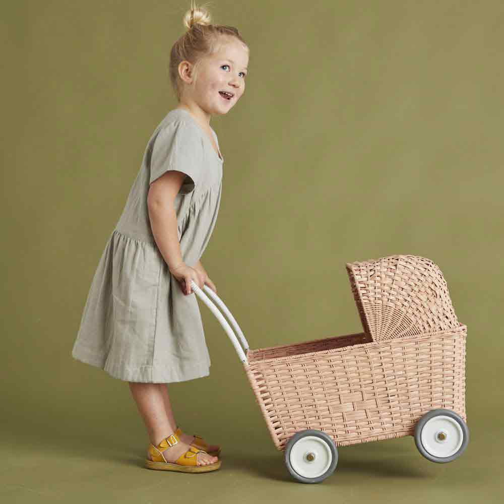 Ein junges Mädchen in einem grauen Kleid und gelben Sandalen lächelt, während es vor einem hellgrünen Hintergrund einen Olli Ella – Puppenwagen Strolley rosa auf Rädern zieht.