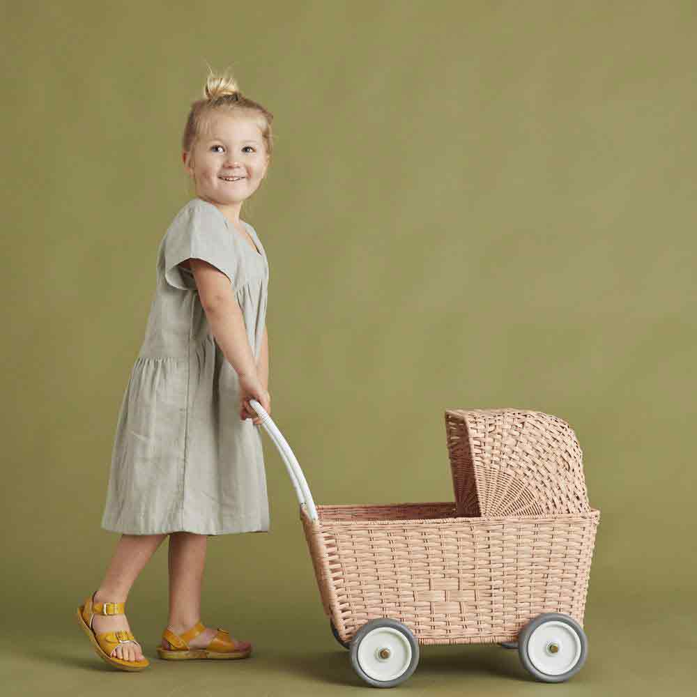 Ein junges Mädchen in einem grauen Kleid und gelben Sandalen lächelt, während es den Griff eines Olli Ella – Puppenwagen Strolley rosa vor einem grünen Hintergrund hält.