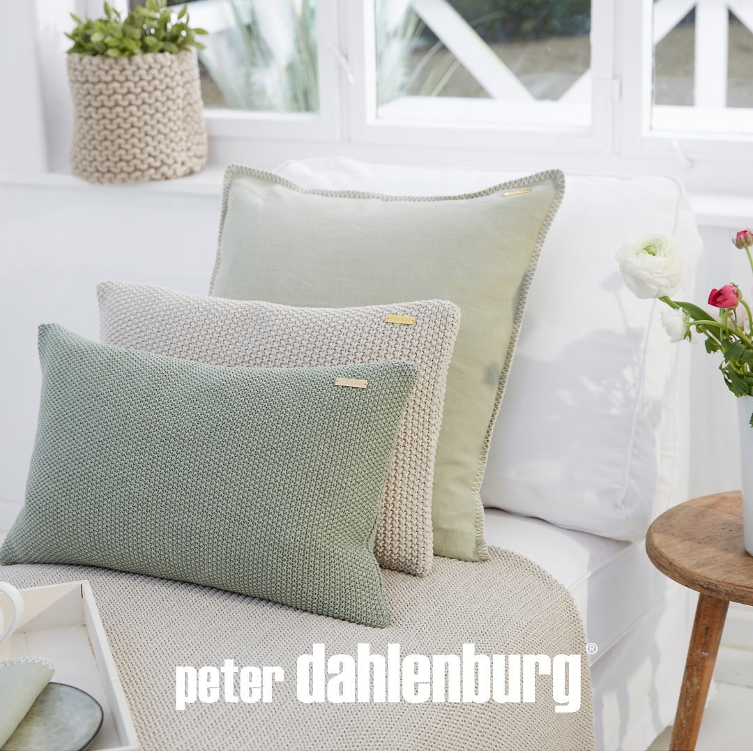 Dekorative Kissen in verschiedenen Texturen und Grün- und Beigetönen, mit einem Markenlogo „Peter Dahlenburg“, arrangiert auf einem weißen Sofa in einem hellen Raum.