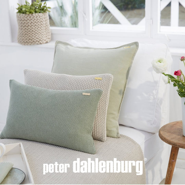 Dekorative Kissen in verschiedenen Texturen und Grün- und Beigetönen, mit einem Markenlogo „Peter Dahlenburg“, arrangiert auf einem weißen Sofa in einem hellen Raum.