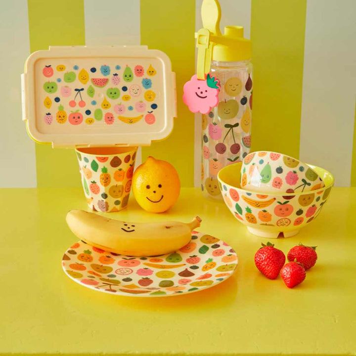Ein farbenfrohes Kindermenüset mit dekorierter Lunchbox, Wasserflasche, Teller, Schüssel und Tasse, präsentiert mit einer lächelnden Banane, Zitrone und Erdbeeren auf einem gelben Tisch.
