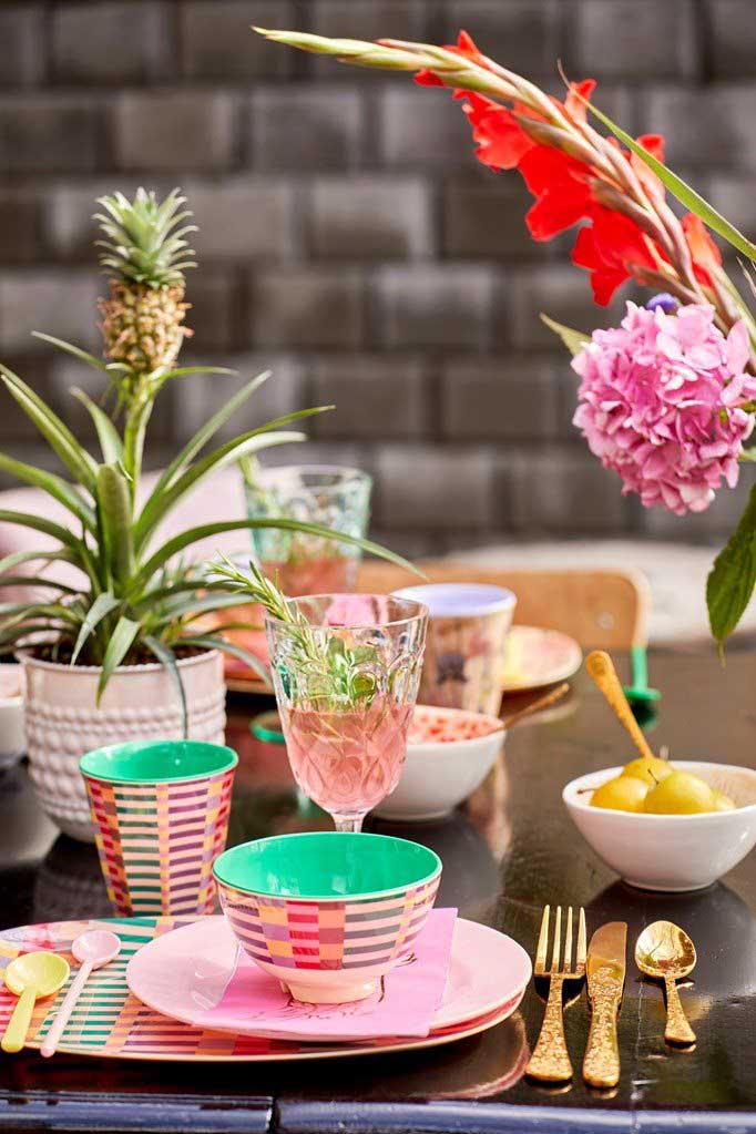 Eine lebendige Essgarnitur für den Außenbereich mit gemustertem Geschirr, goldenem Besteck und einem Tafelaufsatz mit einem Rice-Melaminbecher „Summer Stripes Medium“ und roten Blumen.