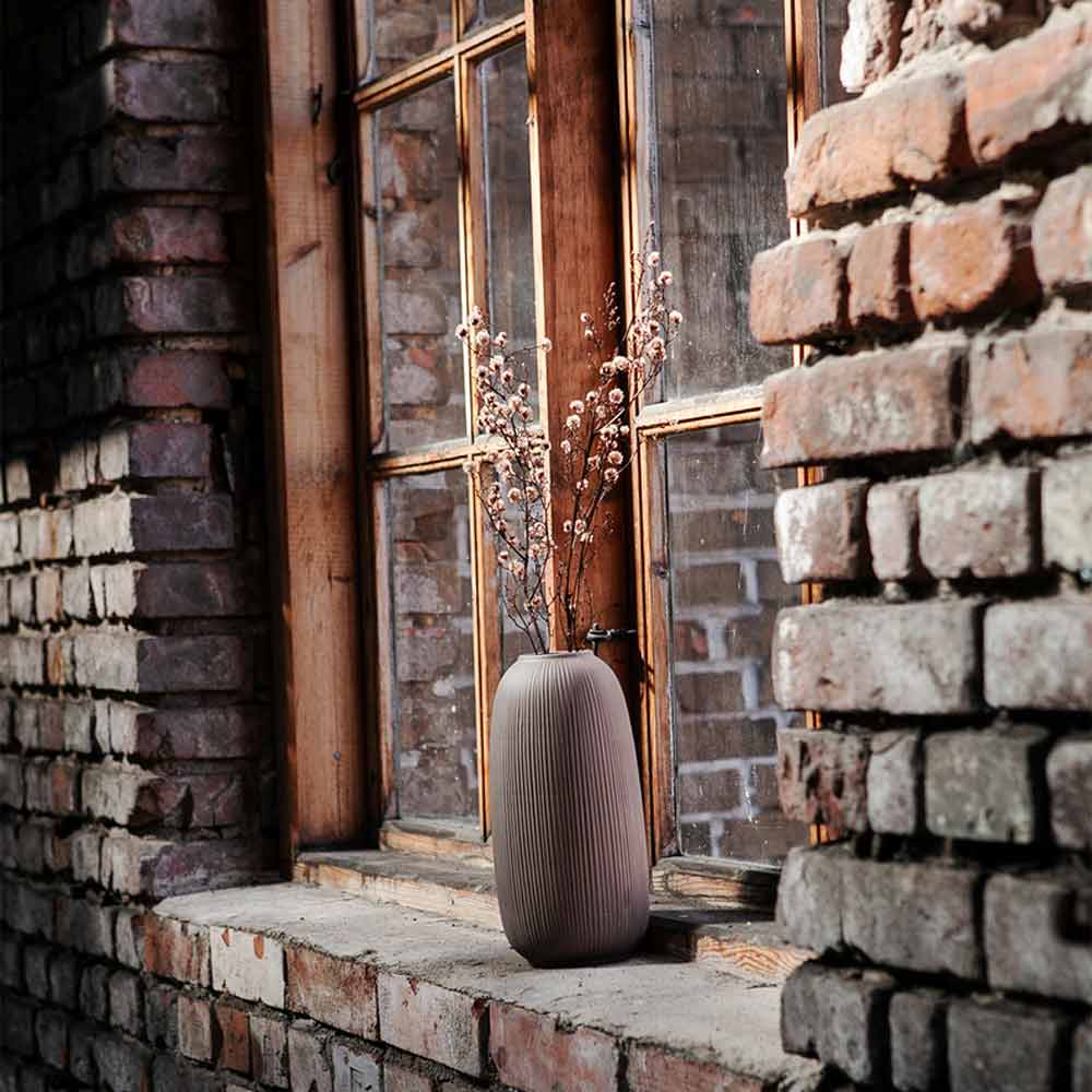 Eine Storefactory – Åby Vase Keramik braun mit getrockneten Blumen auf einer gemauerten Fensterbank neben einem Fenster mit Holzrahmen.
