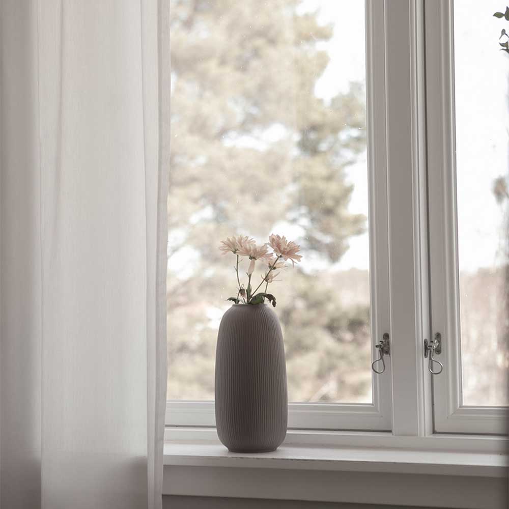 Eine Storefactory – Åby Vase Keramik braun mit Blumen auf einer Fensterbank vor einem Baumhintergrund.