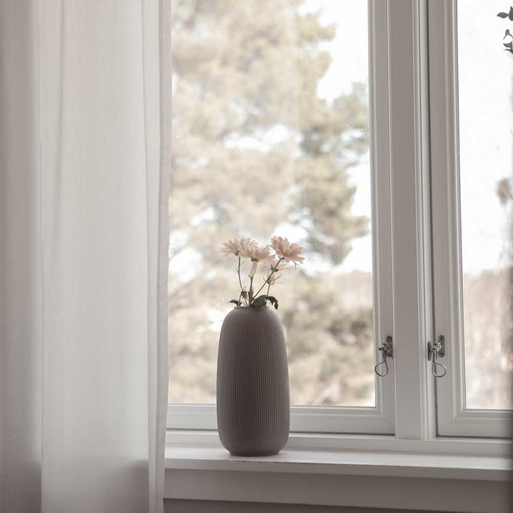 Aufrechte braune gerillte Vase mit wenigen Blüten auf einer weißen Fensterbank neben einem hellen Vorhang