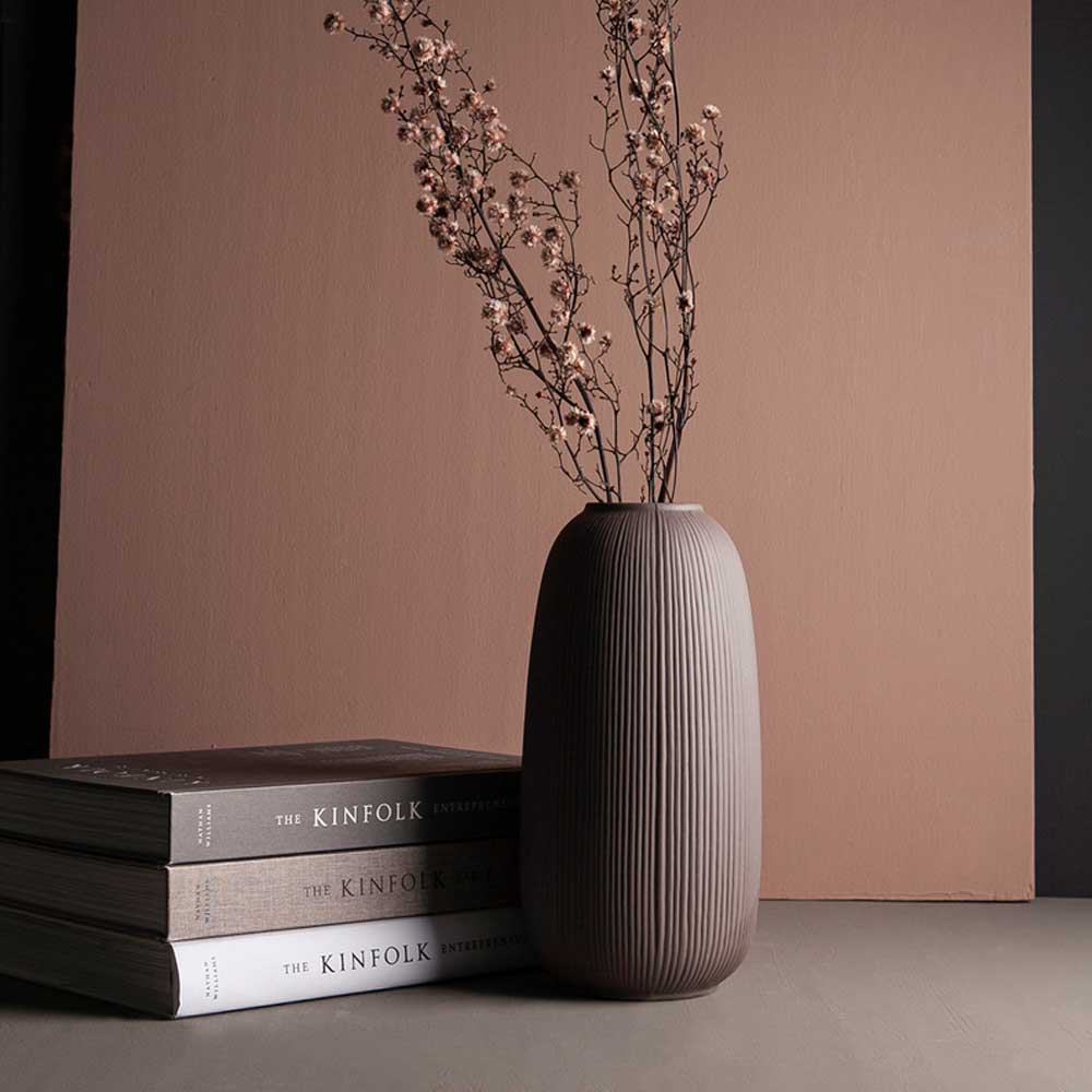 Eine Storefactory – Åby Vase Keramik braun mit getrockneten Zweigen auf einem Tisch neben einem Stapel Verwandtenzeitschriften vor einem zweifarbigen Hintergrund.