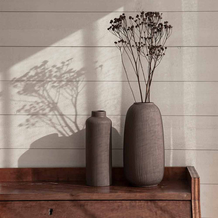 Zwei Storefactory – Åby-Vasen auf einer Holzoberfläche, eine davon mit getrockneten Blumen, die einen Schatten auf eine Wand werfen.