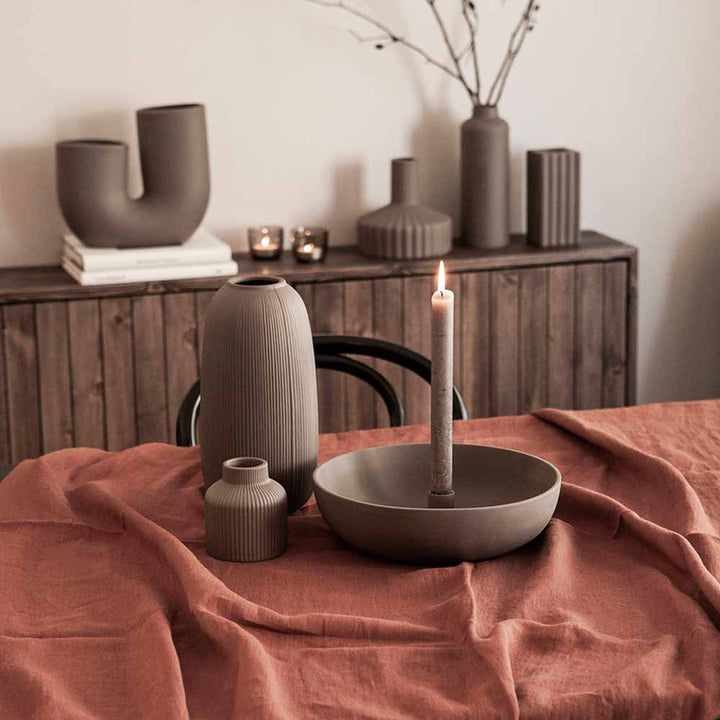 Eine gemütliche Inneneinrichtung mit einer brennenden Kerze und Storefactory - Åby Vase Keramik braun auf einem Tisch, der mit einem Terrakotta-Tuch drapiert ist.
