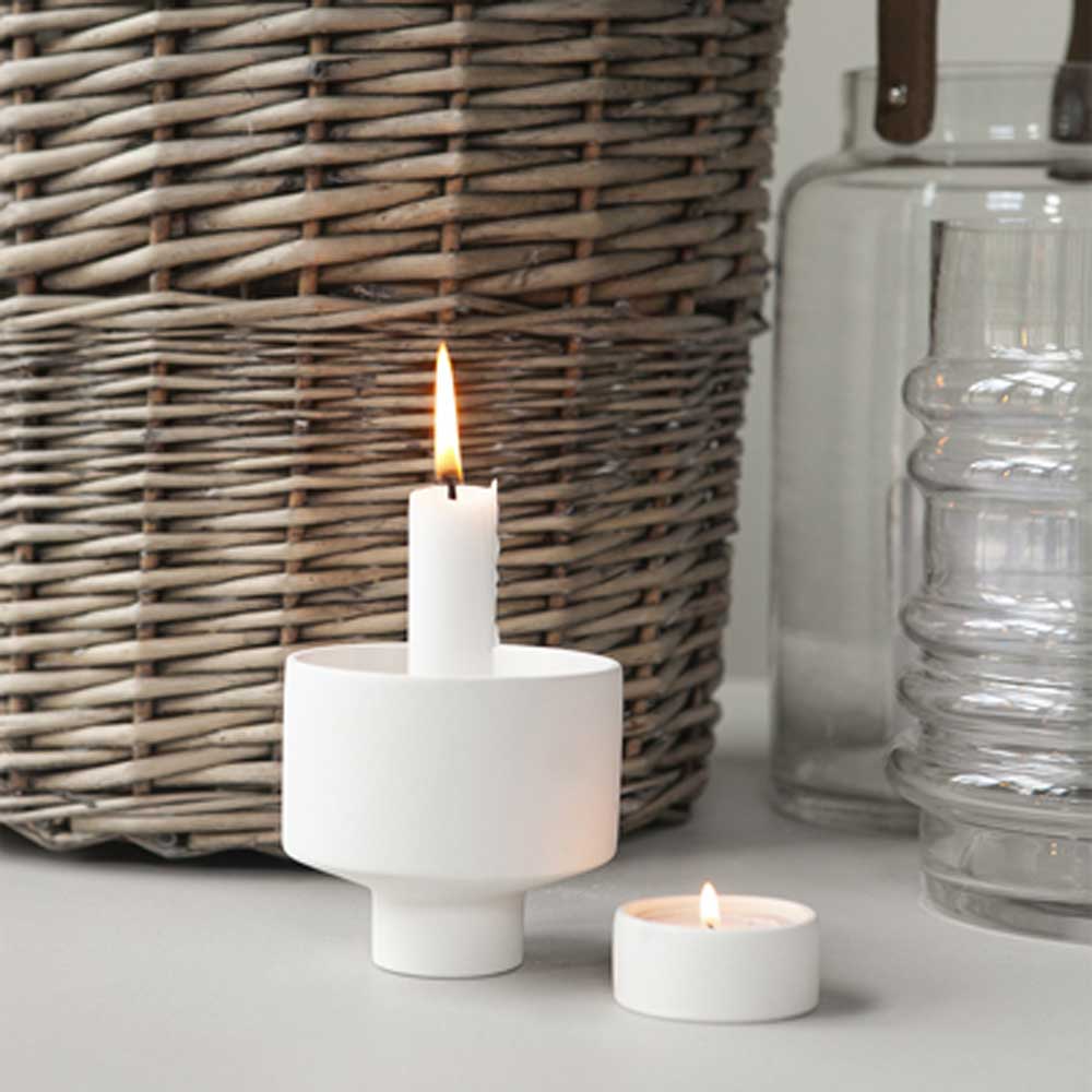 Ein Storefactory - Liaved Kerzenhalter für Stabkerzen und Teelicht weiß steht auf einem Tisch neben einem Weidenkorb.