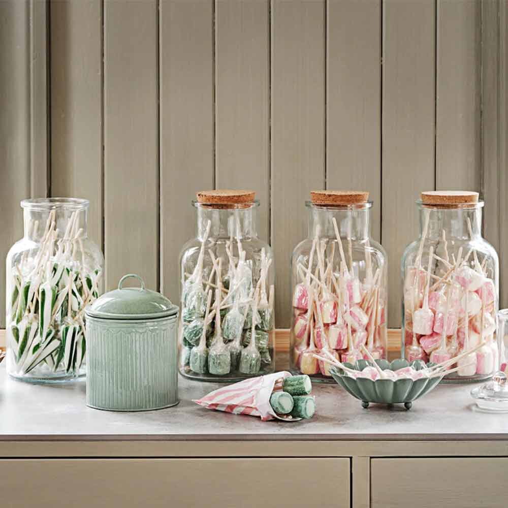 Vier Strömshaga-Lutscher-Vintage-Gläser mit Korkdeckeln, gefüllt mit verschiedenen bunten Süßigkeiten auf einem Holztisch, begleitet von einem grünen Keramikbehälter und einer Schale mit Süßigkeiten.