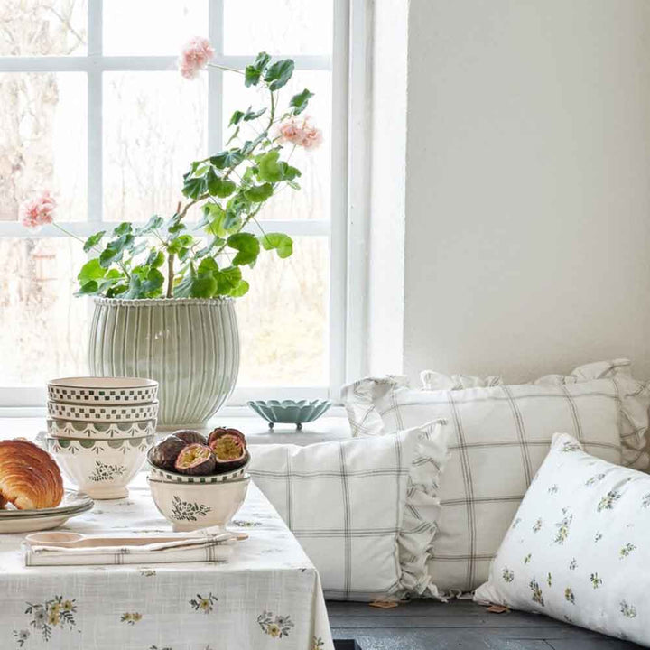 Eine gemütliche Fensterecke mit einem Strömshaga - Kissenbezug Alma 45 x 45 cm, einem Blumentopf, Kissen und einem Frühstücksset.